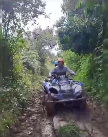 Sentados en una robusta cuatrimoto de color azul, un hombre y una mujer recorren una ruta campestre. Él va como pasajero, abrazándola con confianza, mientras ella pilotea el vehículo todoterreno. Los cascos que portan son esenciales para esta travesía con adrenalina por senderos naturales. A su paso se observa un entorno con abundante vegetación y elevaciones montañosas.